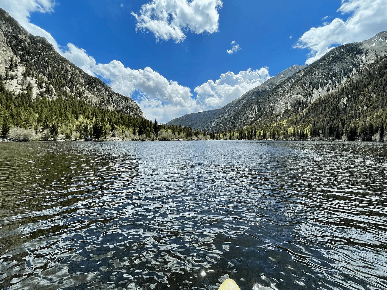 Rippled water on Cottonwood Lake seen from the nose of a paddle board, with pine-covered mountains rising on both sides under a blue sky and white clouds.