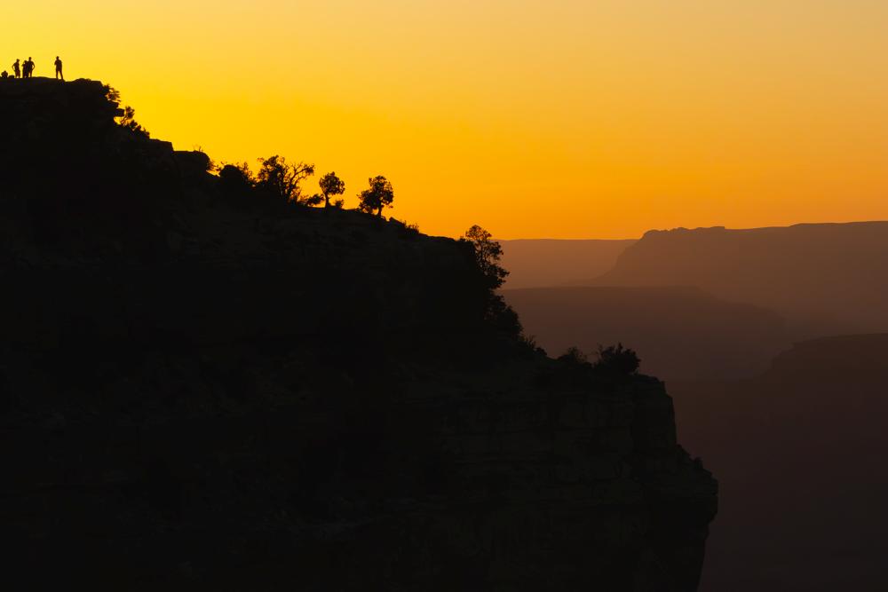 The Grand Canyon rim in silhouette against an orange sunset, with two small figures standing on top of the cliff.