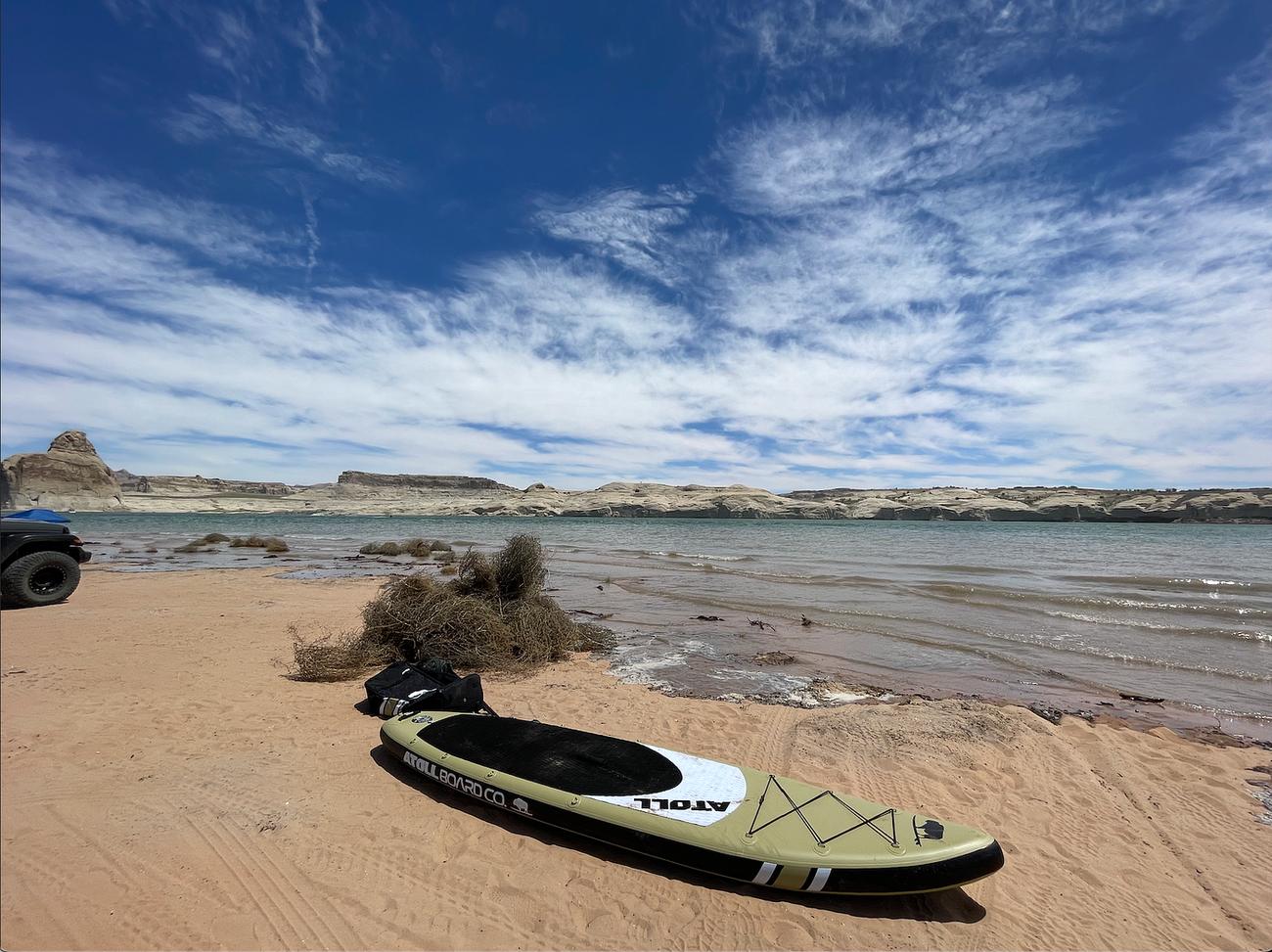 An inflatable paddle board lying on the sand at Lone Rock Beach beside a dry shrub, with the lake and pale sandstone cliffs beyond under streaks of cloud.