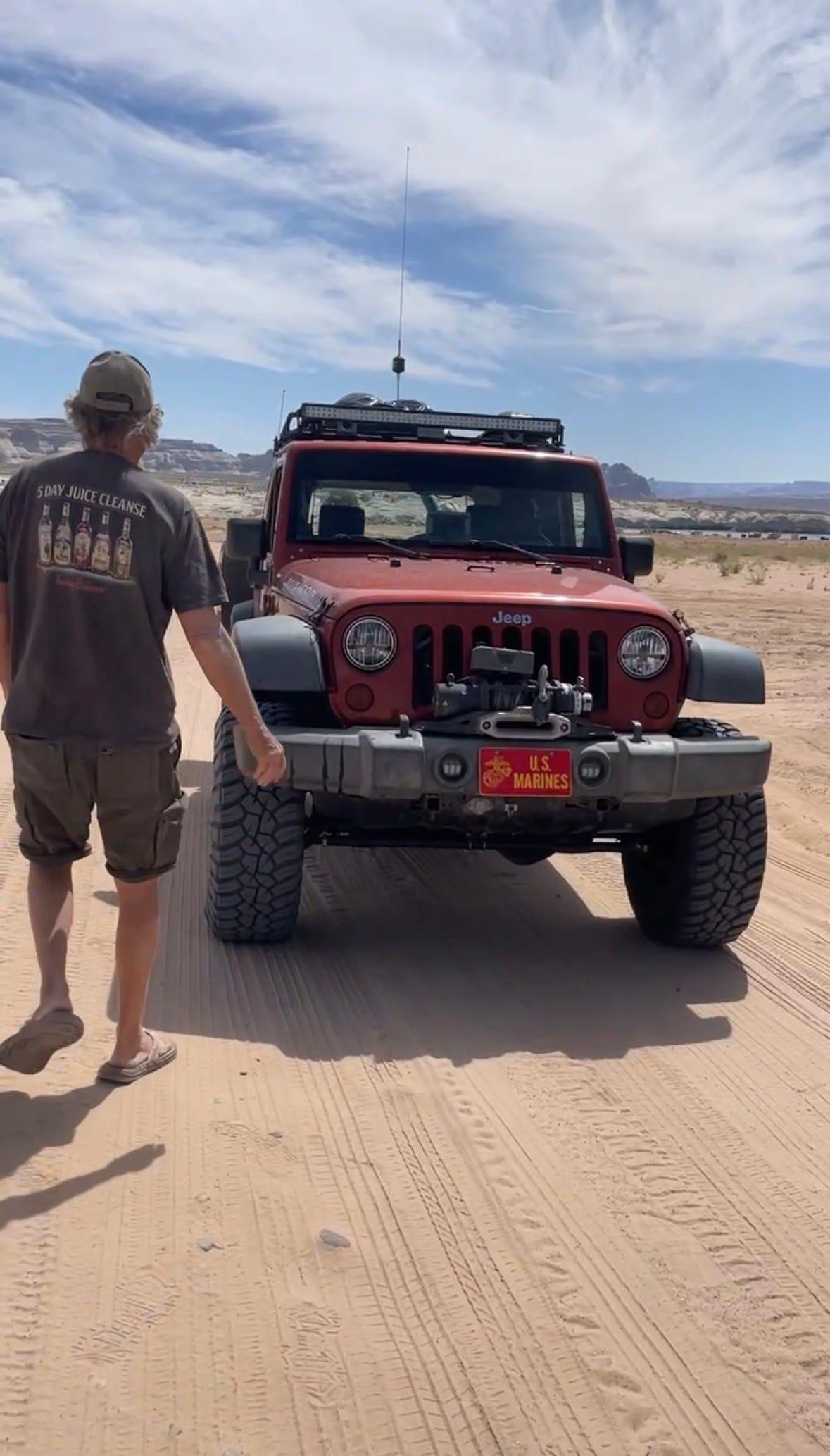 A man in shorts and a cap walks across the sand toward a red Jeep Wrangler parked on a sandy track.