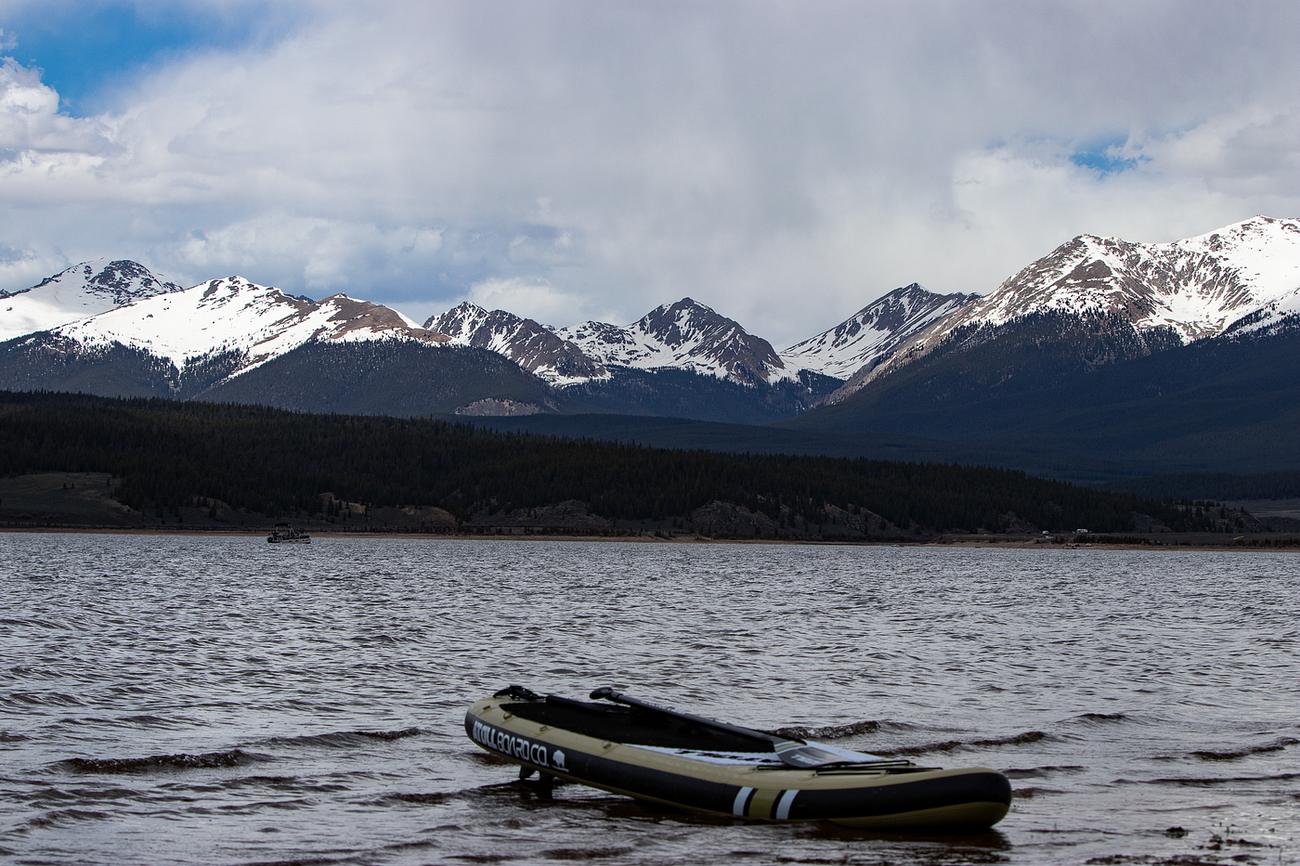 A stand-up paddle board resting in the shallows of Taylor Park Reservoir, with snow-capped peaks beyond a dark, forested shore under a cloudy sky.